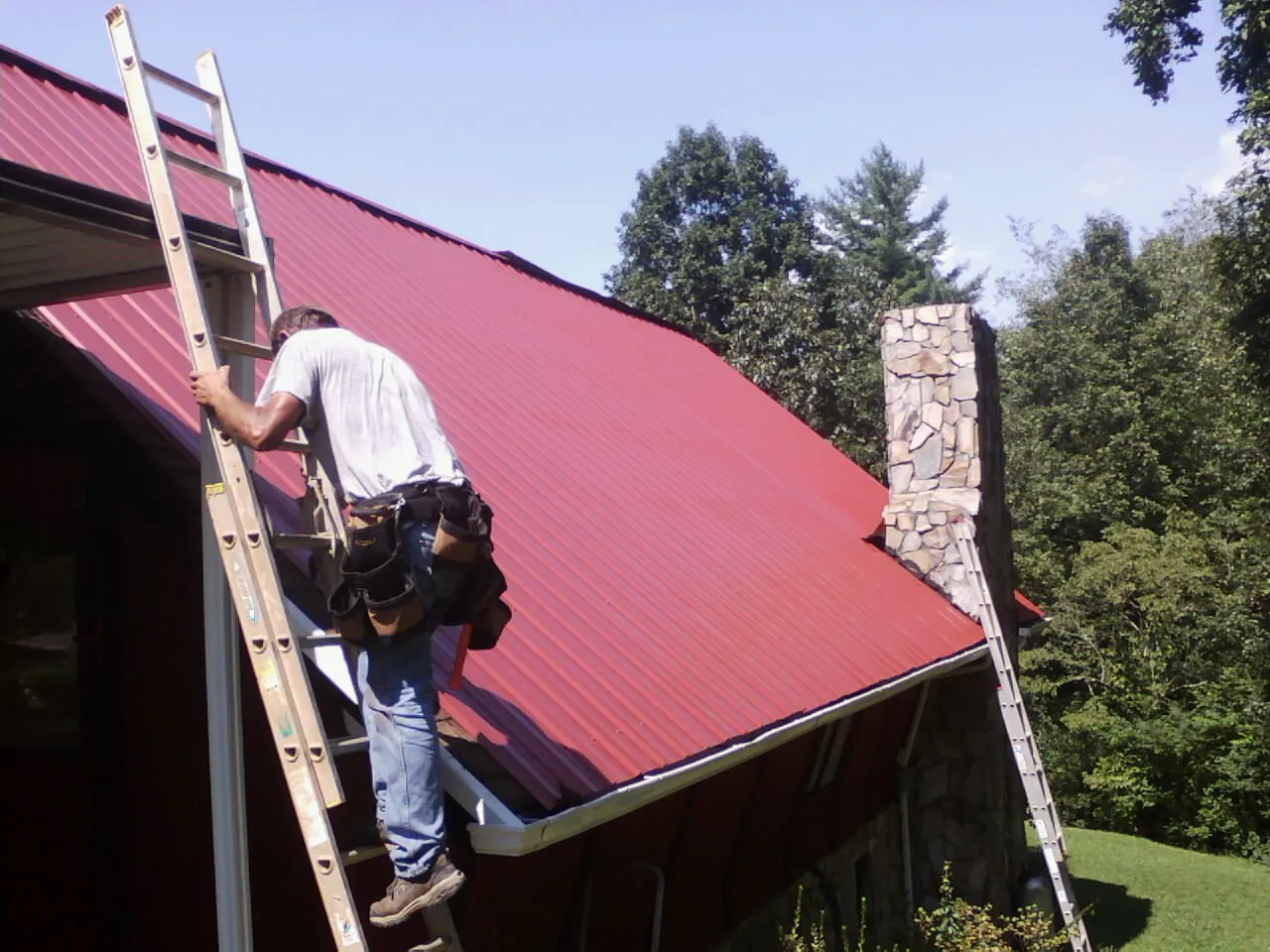 Roofer on a ladder beside a red metal roof and stone chimney — Soffit & Fascia Repair by Roofing Brothers in New Gretna